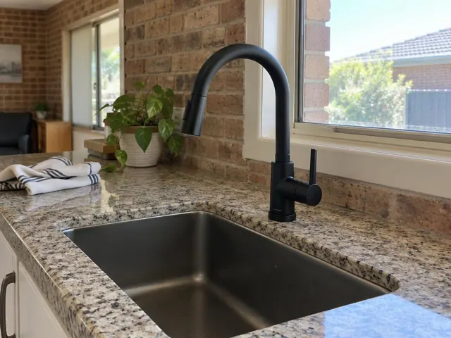 Matte black kitchen mixer tap above a stainless sink on a stone benchtop with brick wall and window in Cliftleigh